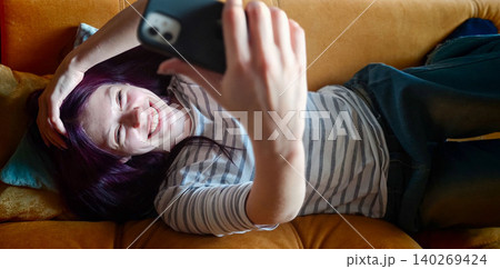 Young woman with purple hair lying on a couch, smiling while taking a selfie with a smartphone, colorful cushions visible in the background 140269424