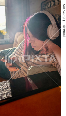 Teenage girl with pink highlights listens to music on smartphone while relaxing on couch at home, wearing white headphones in a sunlit living room Teenage girl with pink highlights listens to music on smartphone while relaxing on couch at home, wearing white headphones in a sunlit living room 140269469