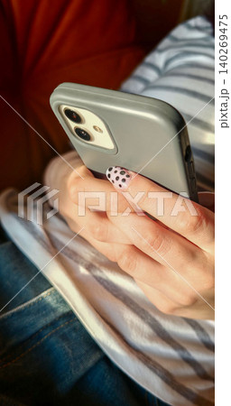 Close-up of a female hand holding a smartphone with a gray case, featuring polka dot nail art, resting on a striped fabric couch in a cozy indoor setting Close-up of a female hand holding a smartphone with a gray case, featuring polka dot nail art, resting on a striped fabric couch in a cozy indoor setting 140269475