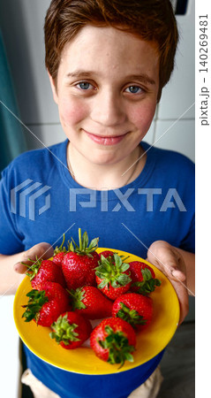 Young boy with brown hair wearing blue shirt holds yellow plate filled with fresh strawberries in a bright kitchen setting with modern decor 140269481