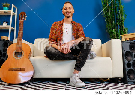 Young caucasian man with dreadlocks sitting on sofa in living room near guitar Young caucasian man with dreadlocks sitting on sofa in living room near guitar 140269894
