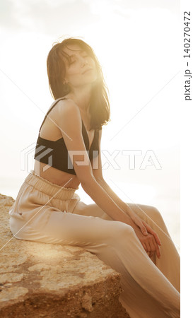 Woman enjoys sunlight while sitting on a rocky surface near the water at sunset Woman enjoys sunlight while sitting on a rocky surface near the water at sunset 140270472