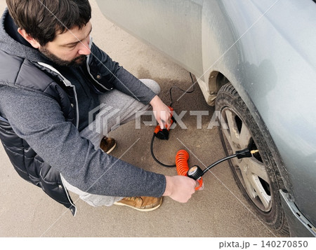 A man inflating a car tire using an electric air compressor . The scene shows routine vehicle maintenance, highlighting practicality and self-reliance. A man inflating a car tire using an electric air compressor . The scene shows routine vehicle maintenance, highlighting practicality and self-reliance. 140270850