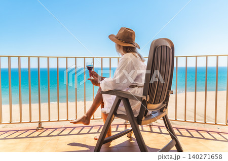 Woman relaxing with glass of red wine on sunny balcony overlooking tropical beach and turquoise sea during summer vacation 140271658