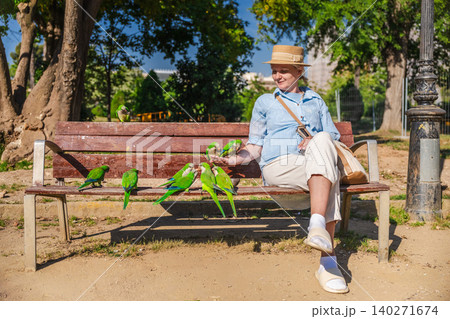 Woman feeding green monk parakeets while relaxing on bench in sunny Barcelona park, tropical urban birds gathering around tourist outdoors Woman feeding green monk parakeets while relaxing on bench in sunny Barcelona park, tropical urban birds gathering around tourist outdoors 140271674