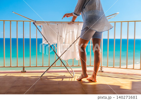 Woman hanging white laundry on drying rack on sunny balcony with turquoise sea view during summer vacation by the beach 140271681