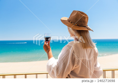 Woman in straw hat holding glass of red wine on balcony with turquoise sea and sandy beach during luxury summer vacation Woman in straw hat holding glass of red wine on balcony with turquoise sea and sandy beach during luxury summer vacation 140271698