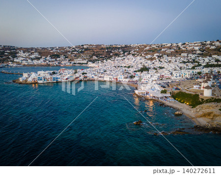A stunning aerial sunset view of Mykonos Old Town, featuring its iconic whitewashed houses, historic windmills, and vibrant port. The golden hues of the Aegean Sea add to the magical island atmosphere 140272611