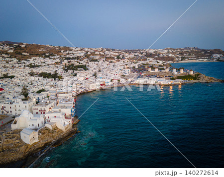 A stunning aerial sunset view of Mykonos Old Town, featuring its iconic whitewashed houses, historic windmills, and vibrant port. The golden hues of the Aegean Sea add to the magical island atmosphere A stunning aerial sunset view of Mykonos Old Town, featuring its iconic whitewashed houses, historic windmills, and vibrant port. The golden hues of the Aegean Sea add to the magical island atmosphere 140272614