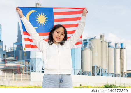Happy asian girl with flag of malaysia standing in front of industrial scenery Happy asian girl with flag of malaysia standing in front of industrial scenery 140272899