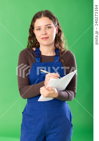 Portrait of girl worker dressed in blue work overalls with papers in hands checking work process standing against green screen Portrait of girl worker dressed in blue work overalls with papers in hands checking work process standing against green screen 140273164