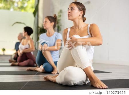 Calm female gym visitor twisting in Ardha Matsyendrasana pose during group yoga training Calm female gym visitor twisting in Ardha Matsyendrasana pose during group yoga training 140273423