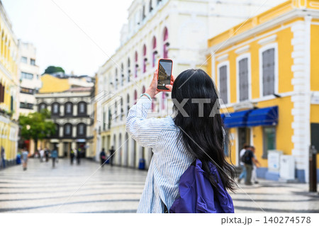 Back view of woman tourist with long hair taking photo with smartphone on beautiful street during solo travel vacation in Macau China 140274578