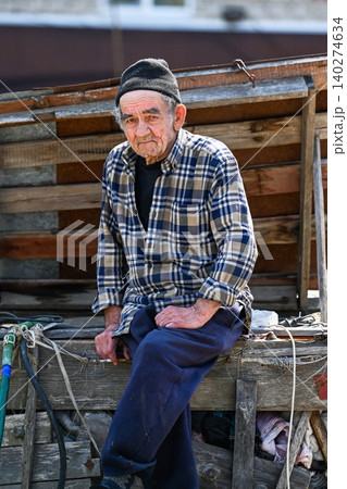Poor russian 77 years old elderly man sits outdoors looking into camera showing concept of poverty in rural village Poor russian 77 years old elderly man sits outdoors looking into camera showing concept of poverty in rural village 140274634