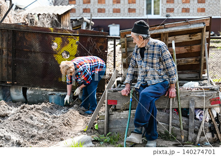 Poor russian senior husband takes break from potato planting while his elderly wife fertilizes soil in rustic countryside garden backyard of village Poor russian senior husband takes break from potato planting while his elderly wife fertilizes soil in rustic countryside garden backyard of village 140274701