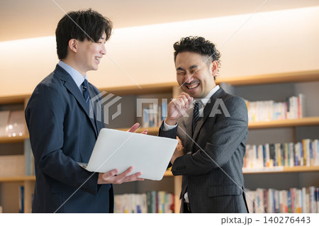 社長室のような本棚の前でパソコンを見ながら打ち合わせをする経営者の上司と部下と笑顔のビジネスマン 社長室のような本棚の前でパソコンを見ながら打ち合わせをする経営者の上司と部下と笑顔のビジネスマン 140276443