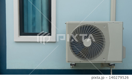 Outdoor air conditioning compressor unit mounted on the exterior wall of a light blue building. A glass window pane is visible next to the ventilation equipment. 140279526