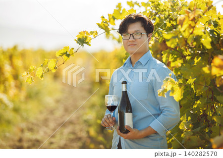 Good looking asian ethnicity sommelier with opened bottle of red wine and poured glass standing on vineyard Good looking asian ethnicity sommelier with opened bottle of red wine and poured glass standing on vineyard 140282570