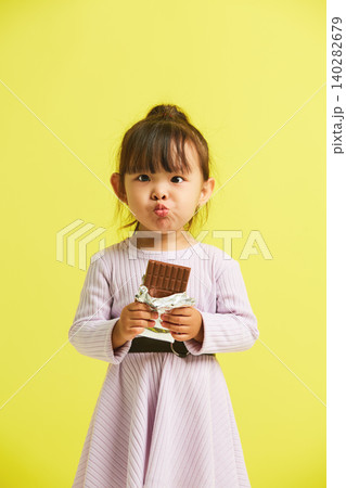 Small female child having addiction to sugar eats bar of chocolate standing over yellow isolated background. Sweet tooth concept. Small female child having addiction to sugar eats bar of chocolate standing over yellow isolated background. Sweet tooth concept. 140282679