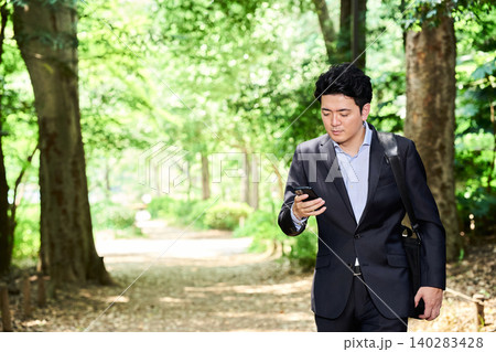 緑豊かな公園でスマートフォンを操作しながら歩く営業の外回りの仕事中の30代の日本人の男性のビジネスマ 緑豊かな公園でスマートフォンを操作しながら歩く営業の外回りの仕事中の30代の日本人の男性のビジネスマ 140283428