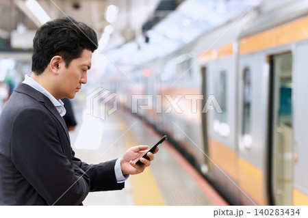 駅のプラットホームで真剣にスマートフォンを操作するスーツ姿で通勤中の30代の日本人の会社員の男性 140283434