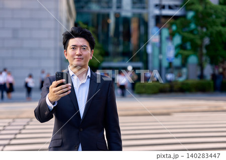 夕方の都会のオフィス街の交差点でスマートフォンを持って街中に立つ30代の日本人の男性会社員 140283447