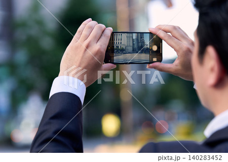 都会の街並みをスマートフォンで撮影するスーツ姿の会社員の男性の顔無しの後ろ姿 140283452