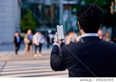 都会の帰宅ラッシュ時にの交差点でスマートフォンを持ち画面を見て操作する顔無しの会社員の男性の後ろ姿 140283454