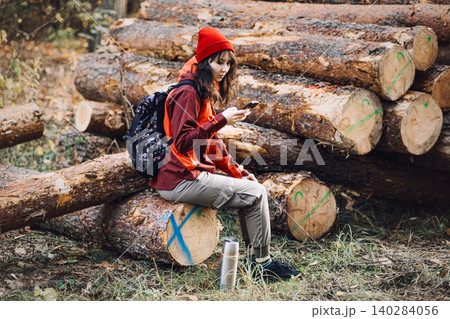Woman sits on log in forest and looks at smartphone screen. Smartphone usage in forest, mobile roaming for tourists, digital navigation tools. 140284056