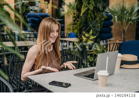 Young woman gestures and smiles while using laptop at outdoor cafe table. Female mentor teaching online, virtual education instructor, educational content creator. Young woman gestures and smiles while using laptop at outdoor cafe table. Female mentor teaching online, virtual education instructor, educational content creator. 140284118