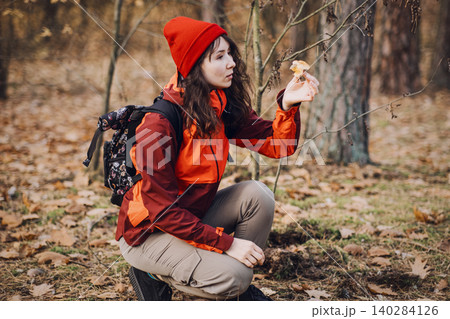 Woman crouches in forest and examines small wild mushroom. outdoor survival techniques, primitive skills training, wilderness self-reliance, bushcraft foraging guide. Woman crouches in forest and examines small wild mushroom. outdoor survival techniques, primitive skills training, wilderness self-reliance, bushcraft foraging guide. 140284126