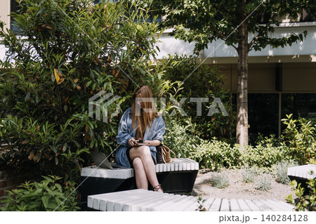 Woman sits on bench in green courtyard writing in notebook next to laptop computer. Connected work lifestyle, mobile technology productivity, healthy workspace choice. 140284128