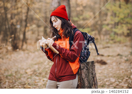 Woman stands in forest while writing in notebook and leaning on tree stump. Nature journaling therapy, mindfulness forest walking, mental health recovery. Woman stands in forest while writing in notebook and leaning on tree stump. Nature journaling therapy, mindfulness forest walking, mental health recovery. 140284152