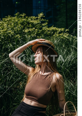 Woman sits outdoors with closed eyes holding straw hat in front of green plants. Nature integrated urban design, bio living walls concept, urban agriculture for cooling Woman sits outdoors with closed eyes holding straw hat in front of green plants. Nature integrated urban design, bio living walls concept, urban agriculture for cooling 140284158