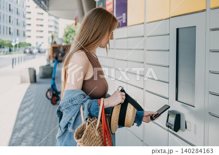 Woman presents smartphone to automated parcel station scanner while holding bags and straw hat. Click and collect retail service, online shopping parcel pickup, consumer convenience economy lifestyle. Woman presents smartphone to automated parcel station scanner while holding bags and straw hat. Click and collect retail service, online shopping parcel pickup, consumer convenience economy lifestyle. 140284163