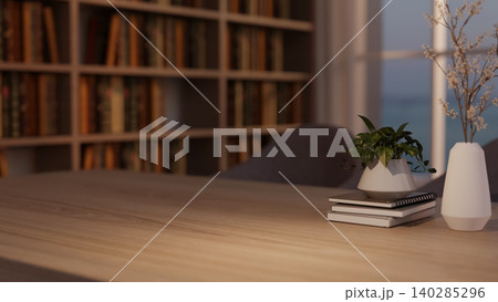 A wooden table with potted plant on books and flower vase across bookshelf and window in dim library 140285296