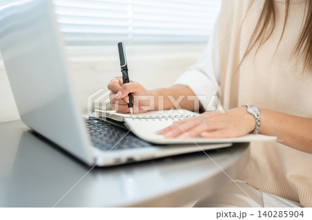 Close up of woman's hand holding pen writing in notebook over laptop while sitting at table in cafe 140285904