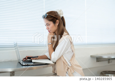 Asian woman resting head on hand and looking at laptop while sitting at table in cafe or home office Asian woman resting head on hand and looking at laptop while sitting at table in cafe or home office 140285905