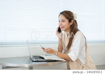 Happy asian woman talking on phone holding credit card over laptop at table in cafe or home office. Happy asian woman talking on phone holding credit card over laptop at table in cafe or home office. 140285911