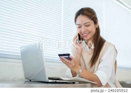 Happy asian woman talking on phone looking at credit card over laptop at table in cafe home office. Happy asian woman talking on phone looking at credit card over laptop at table in cafe home office. 140285912