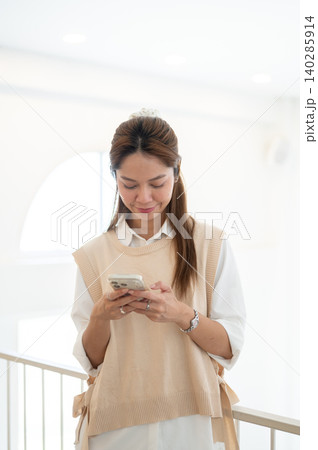 Happy asian woman texting looking at phone while standing leaning against railing in cafe or at home Happy asian woman texting looking at phone while standing leaning against railing in cafe or at home 140285914