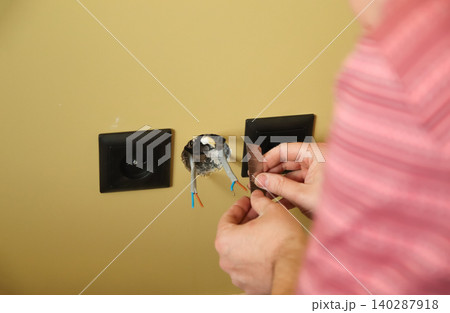 Close-up of electrician hands stripping electrical wires for socket installation. Electrician using utility knife blade to repair power outlet in a yellow wall, domestic home renovation concept 140287918