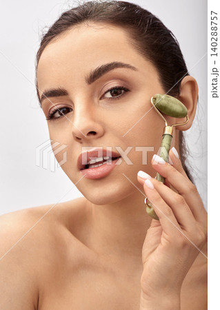 Facial jade roller. Young woman massages her sensuality face using a green jade massager roller for elastic perfect skin 140288757