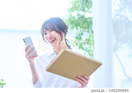荷物を受け取りスマホで確認して笑う窓辺の女性 荷物を受け取りスマホで確認して笑う窓辺の女性 140289084