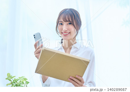 明るい窓辺で荷物とスマホを手に微笑む若い女性 140289087