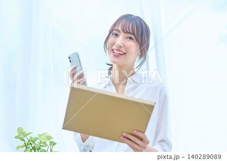 荷物を抱えスマホを手に笑顔で見つめる窓辺の女性 荷物を抱えスマホを手に笑顔で見つめる窓辺の女性 140289089