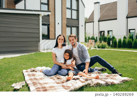 Happy heteroseksual family sitting on grass near their new house. Family sitting on lawn, buying new big home . Happy heteroseksual family sitting on grass near their new house. Family sitting on lawn, buying new big home . 140289269