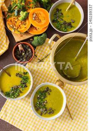 A table displays bowls of pumpkin soup with broccoli topped with seeds and herbs. Nearby, fresh ingredients and a pot hold more soup, creating a healthy meal setting A table displays bowls of pumpkin soup with broccoli topped with seeds and herbs. Nearby, fresh ingredients and a pot hold more soup, creating a healthy meal setting 140289337