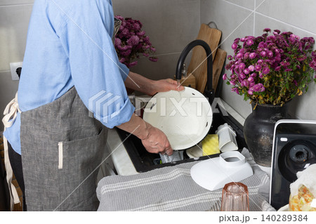 A man stands at the sink in his kitchen, cleaning dirty dishes and utensils. Flowers are placed on the counter, adding color to the scene in daylight A man stands at the sink in his kitchen, cleaning dirty dishes and utensils. Flowers are placed on the counter, adding color to the scene in daylight 140289384