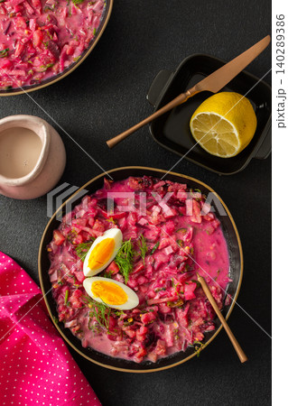 A vibrant bowl of cold beetroot soup features hard-boiled eggs and fresh herbs. A lemon slice accompanies it, all set against a black surface with a pink cloth for contrast A vibrant bowl of cold beetroot soup features hard-boiled eggs and fresh herbs. A lemon slice accompanies it, all set against a black surface with a pink cloth for contrast 140289386